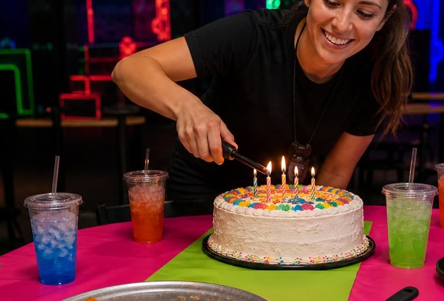 Laserdome party host lighting birthday cake candles at a bright party table with pizza, colorful sodas, and a cake server